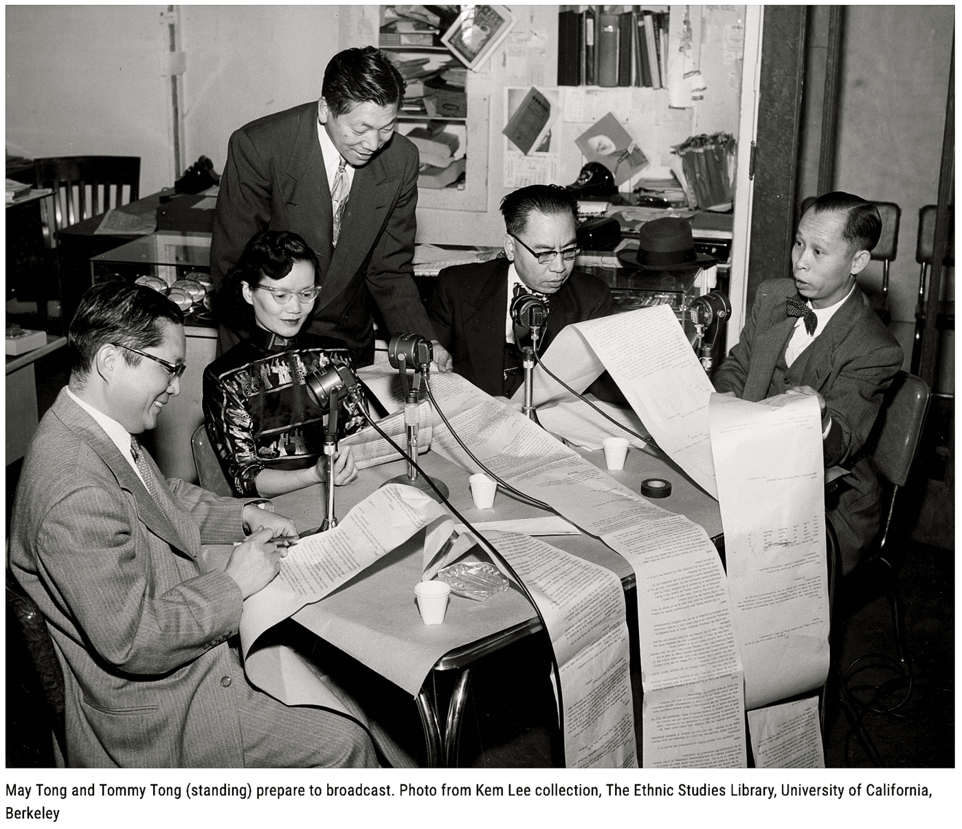 May And Tommy Tong As They Prepare To Broadcast. Image Of Four People Sitting Around A Table And One Man Standing Over The Woman Looking At Long Rolls Of Paper.
